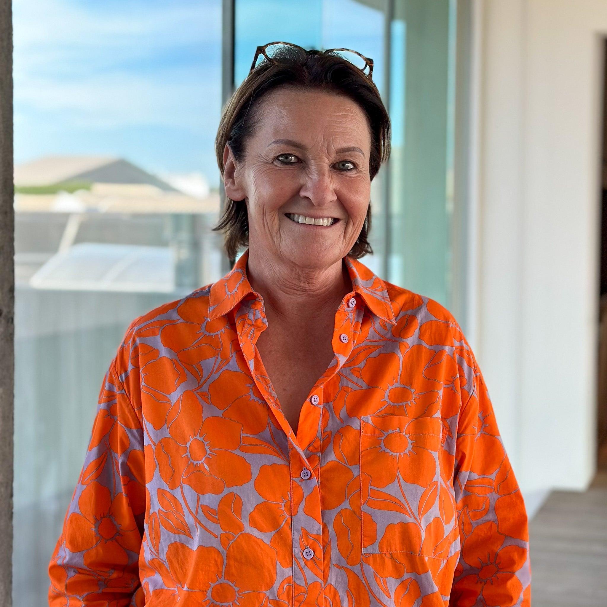 Ann Warrot smiling, wearing a vibrant orange shirt with grey floral patterns, glasses on her head.