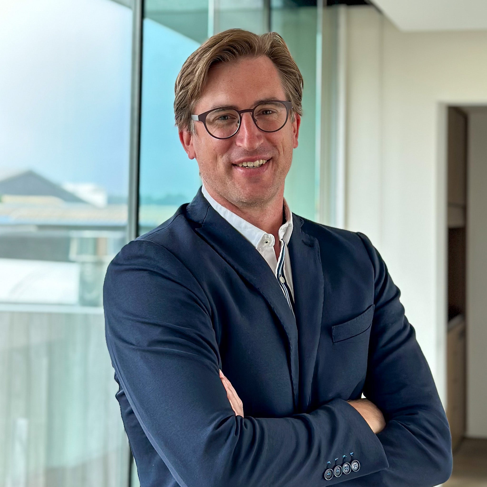 Smiling man, Jochen Demets, in a navy suit and glasses, arms crossed, in a modern office.