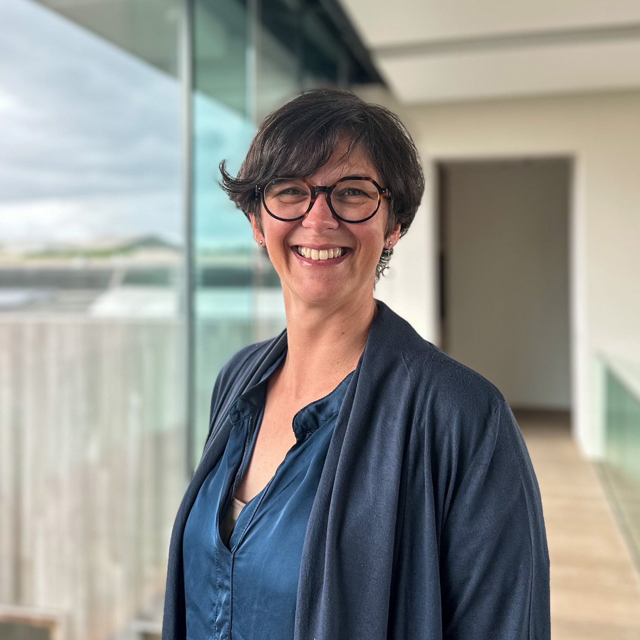 Smiling woman with short dark hair and tortoiseshell glasses, wearing a blue top and dark cardigan.