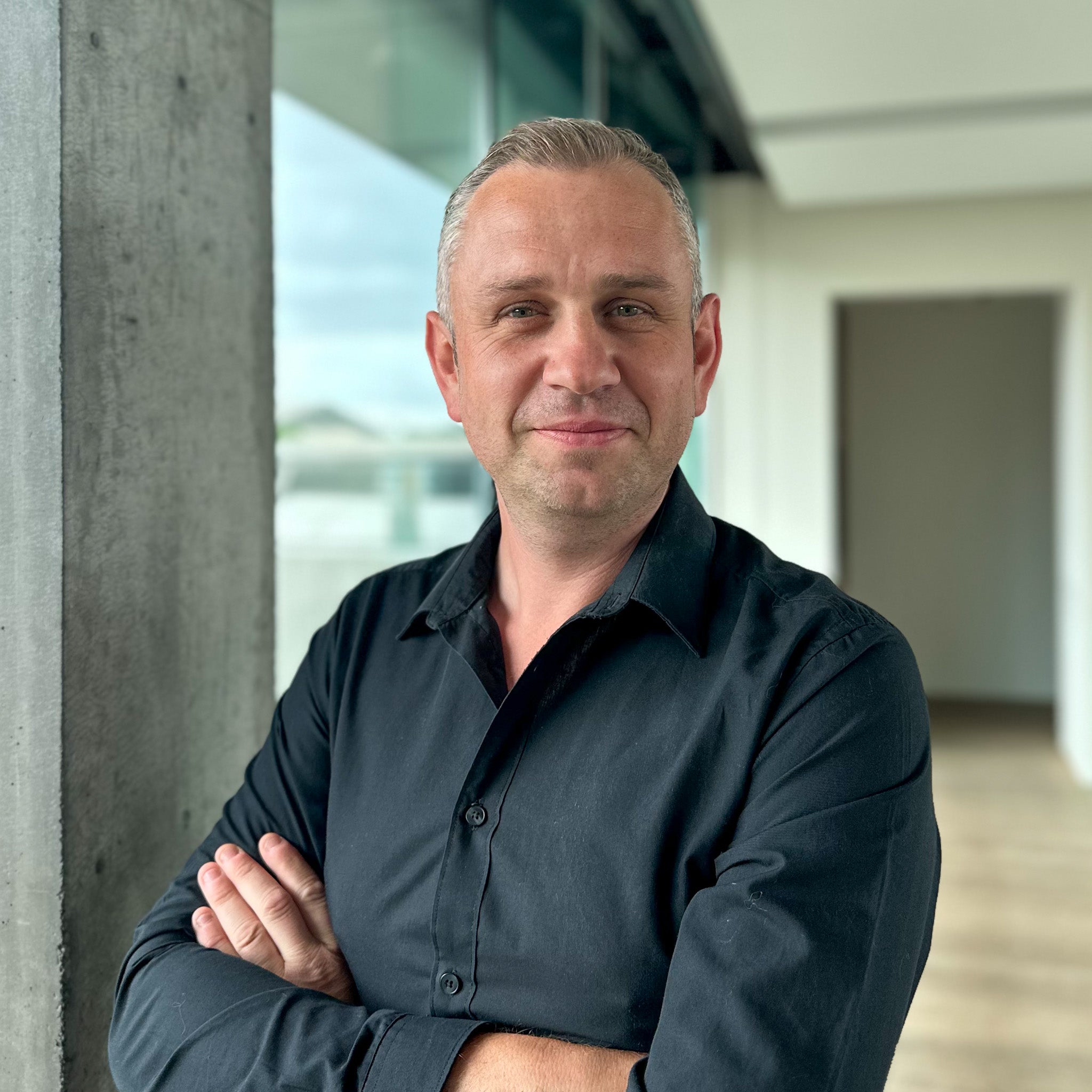 Michael De Jaegere, a man with short gray hair, wearing a black shirt, arms crossed, smiling.